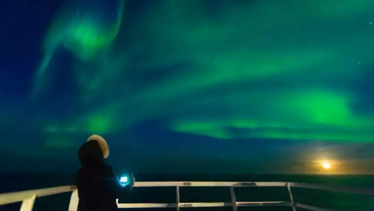 Arctic traveler stands on deck of a small ship at night, photographing wavy green northern lights as the sun sets on the horizon.
