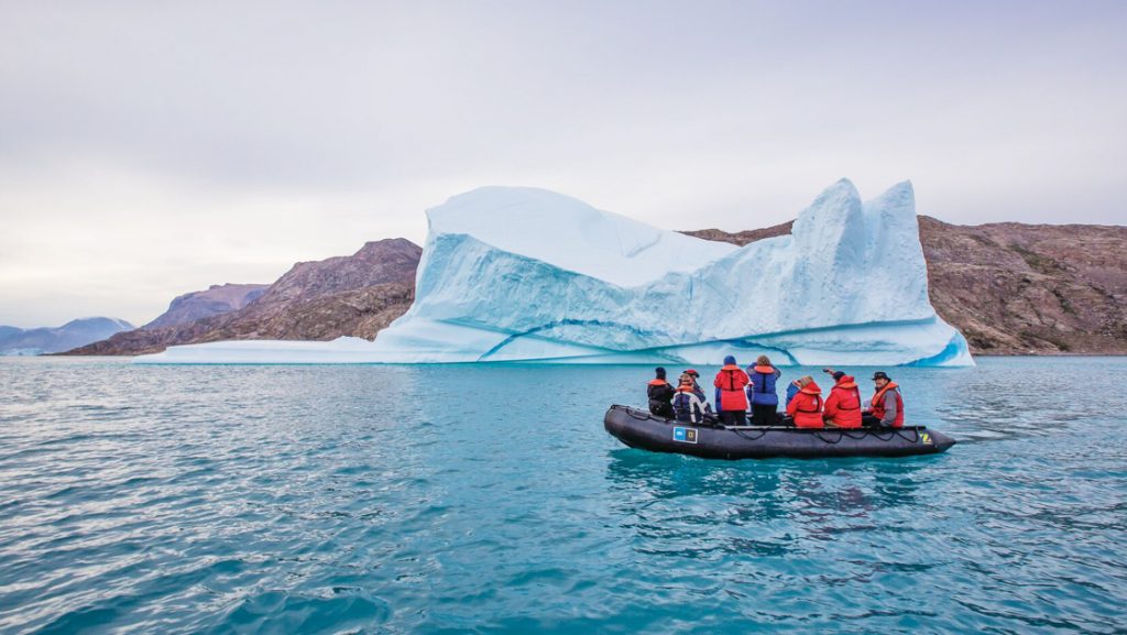Black inflatable Zodiac dinghy with Arctic travelers motors past a large iceberg in calm water on an Iceland to Greenland cruise,