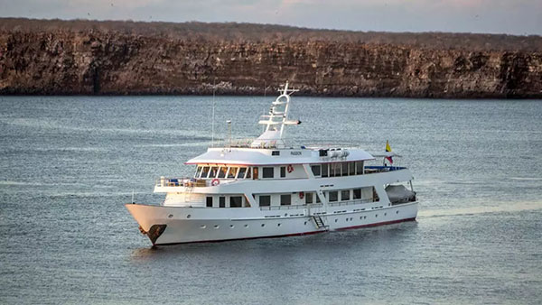 Galapagos cruise ship Passion is seen in the water by a brown, rocky bluff shoreline sailing toward the camera from its port side