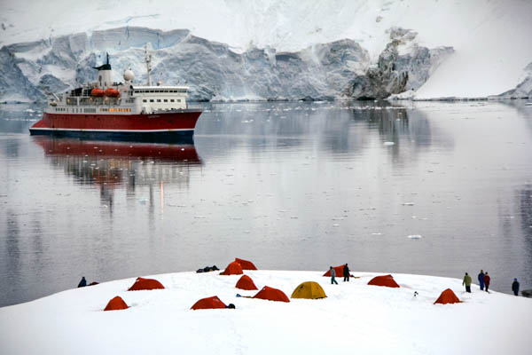 Tents seen on a snowy hill with an Antarctic expedition ship and glacier in the background