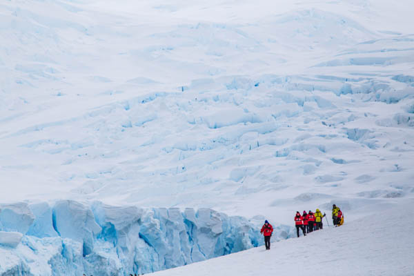 Six Antarctica cruise passengers in red and yellow jackets stand near a glacier.