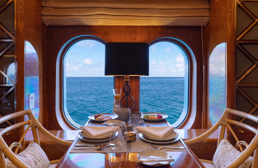 Private breakfast table set for 2 with woven chairs in wooden ship with 2 oval windows looking onto the sea on a romantic cruise.