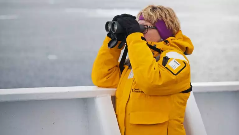 Woman in yellow parka stands on deck of ship looking through binoculars on an Arctic Saga: Spitsbergen, Faroes & Jan Mayen cruise.