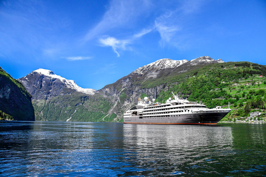 Small expedition ship with gray hull & white upper decks cruises in glassy water by lush green mountains with snowy peaks.