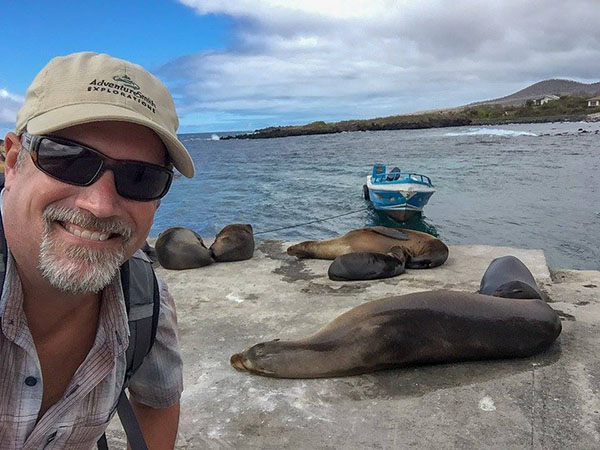 A man in sunglasses and a tan AdventureSmith hat smiles in from of Galapagos sea lions by the shore