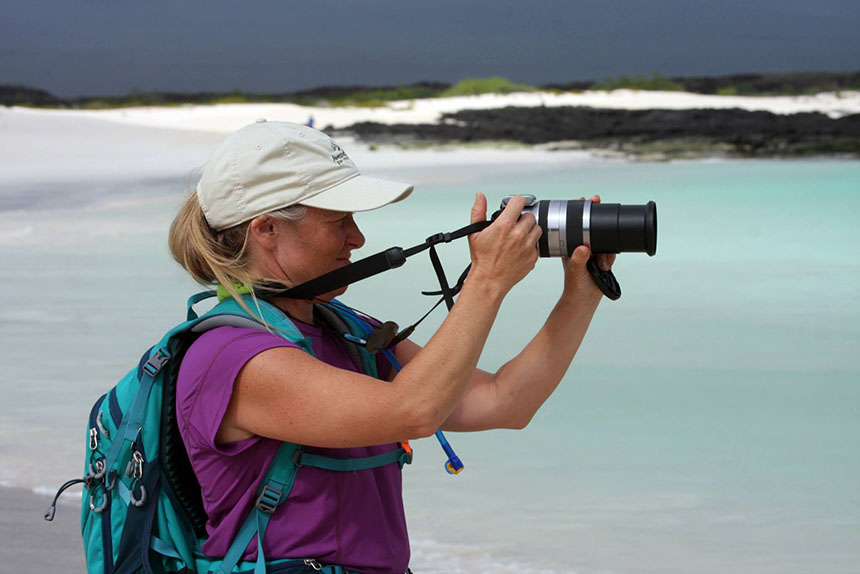 A female traveler in tan hat and purple shirt seen from the side pointing her camera with turquoise water and a white sand shoreline behind her