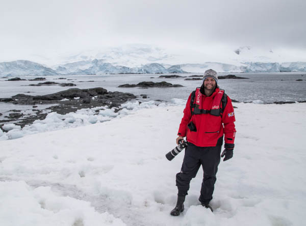 A male traveler in a red jacket holding a large camera at his side smiles amid snow and ice near the shore in Antarctica