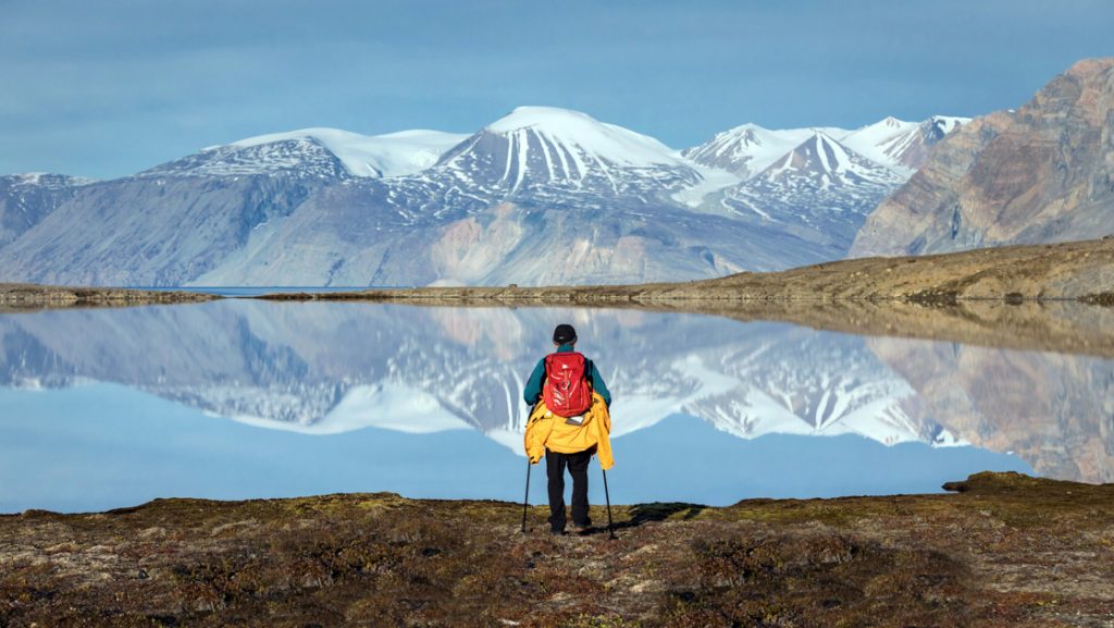 Arctic traveler with yellow coat & red backpack stands at edge of glassy alpine lake with snow-covered peaks in distance.