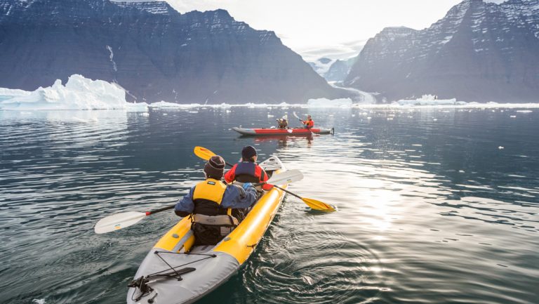2 inflatable tandem kayaks are paddled by travelers through calm water by mountains & icebergs on a Four Arctic Islands cruise.