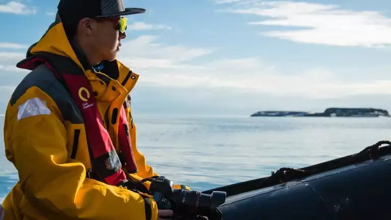 Man in yellow coat, bright sport sunglasses & dark ballcap sits with camera in Zodiac boat, cruising in calm Arctic water.
