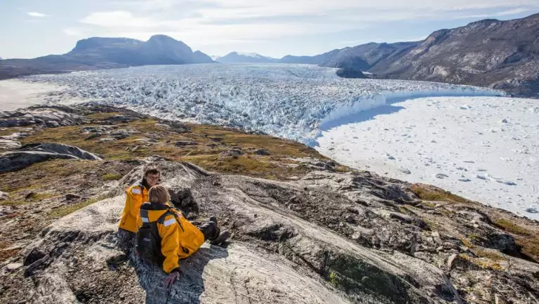 2 women Arctic travelers in yellow parkas sit atop a mountain of rock & tundra overlooking a large glacier in west Greenland.
