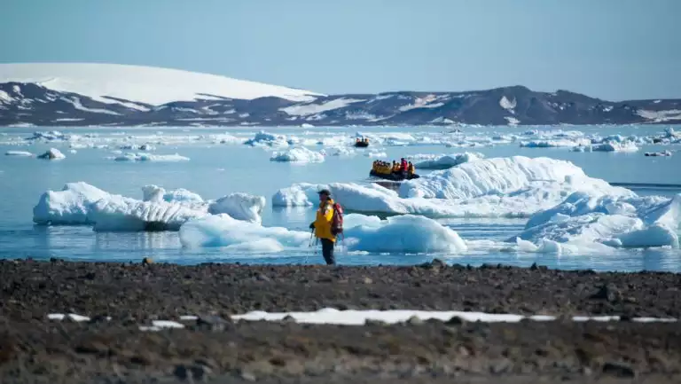 Arctic traveler in yellow parka stands on rocky shore as Zodiacs with others cruise around an icy bay on a west Greenland cruise.