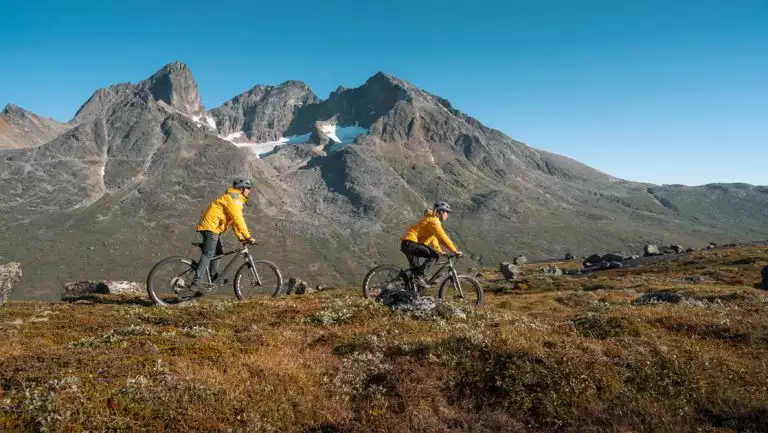 2 Gems of West Greenland cruise travelers in yellow coats ride mountain bikes over arctic tundra by mountains on a sunny day.