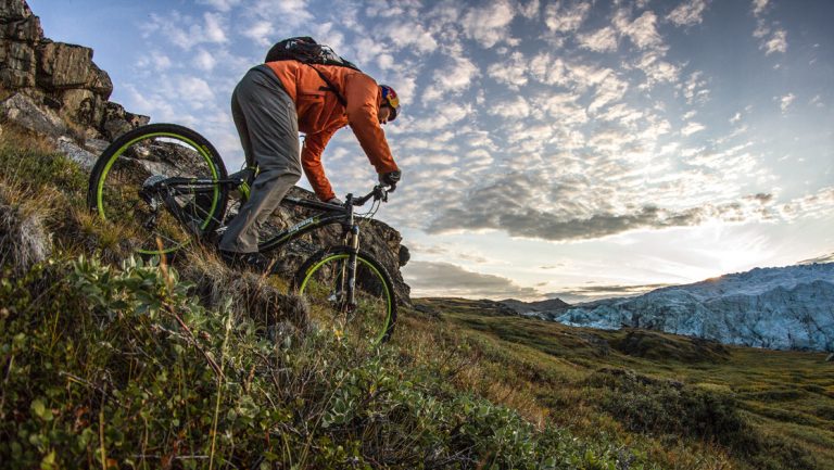 Mountain biker rides down over green tundra on a single track near Russell Glacier during the Gems of West Greenland cruise.