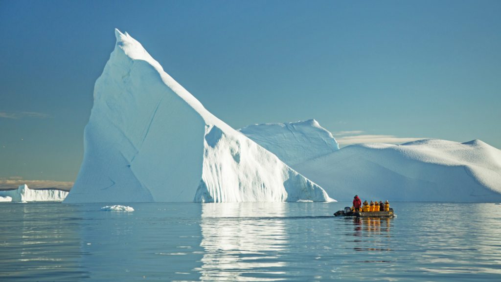 Zodiac with west Greenland travelers motors past massive pointed iceberg rising out of calm water on a sunny day.