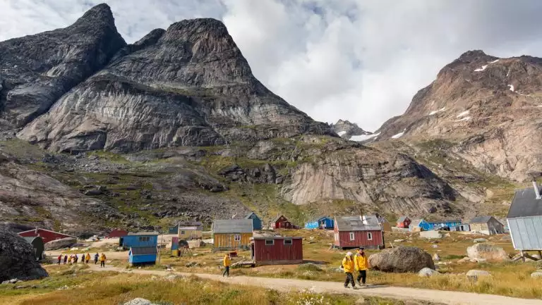 Small group of Greenland Explorer cruise travelers walk a dirt road beside colorful & weathered homes in a remote village.
