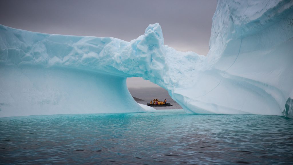 Zodiac with Greenland Explorer cruise travelers peeks through a hole in an a large blue iceberg in calm sea under cloudy skies.