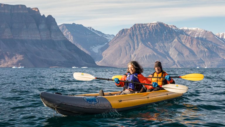 Tandem kayakers paddle a yellow inflatable boat in calm water with brown mountains behind on the Greenland Explorer cruise.