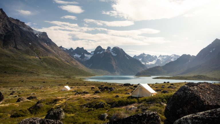 2 white canvas tents sit in Arctic tundra by calm fjord with snowcapped peaks, seen on the Greenland Explorer cruise.