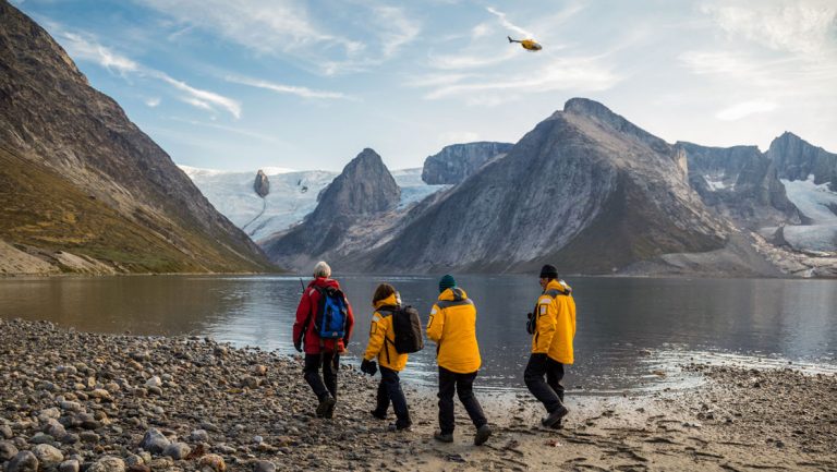 Arctic travelers in yellow or red parka walk a rocky beach by a fjord with a glacier, mountains & helicopter in the distance.