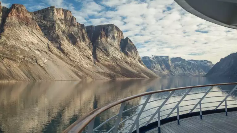 View from empty ship deck with wraparound railing & teak decking, in glassy fjord with tan cliff bands under clouds.