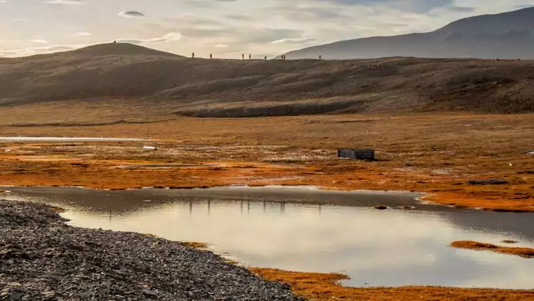 Northwest Passage cruise guests walk a ridgeline on a sloping mountain overlooking autumnal tundra & a small pond.