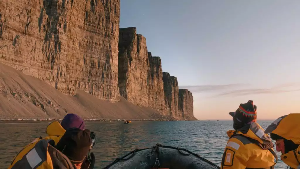 3 Northwest Passage cruise guests in yellow coats ride in a Zodiac boat along tall tan cliffs at sunset.