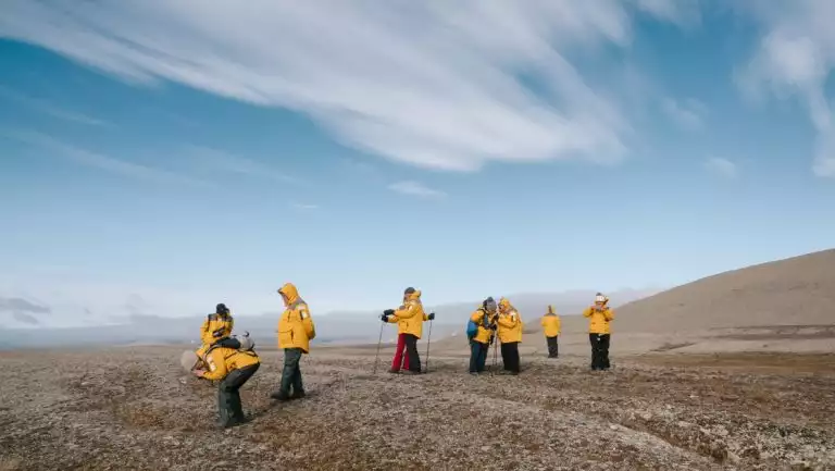 Small group of Northwest Passage cruise guests in yellow jackets walk over brown tundra & take photos on a sunny day.