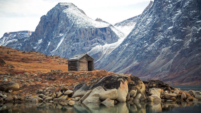 Small historic hut stands at water's edge above a shore covered in boulders with snowy peaks behind in the Northwest Passage.