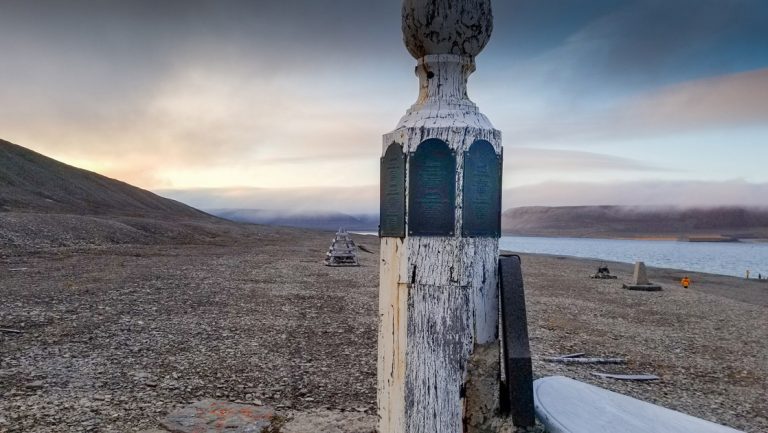 Aged white wood pillar monument with weathered metal plaques sits on rocky shoreline on a northwest passage small ship cruise.