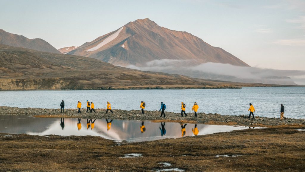 Line of Northwest Passage small ship cruise travelers in yellow parkas walks between the sea & a small pond with mountains behind.