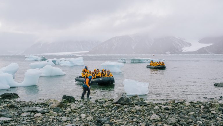 Travelers on a Northwest Passage small ship cruise come into a rocky shoreline by Zodiac, past iceberg bits under clouds.