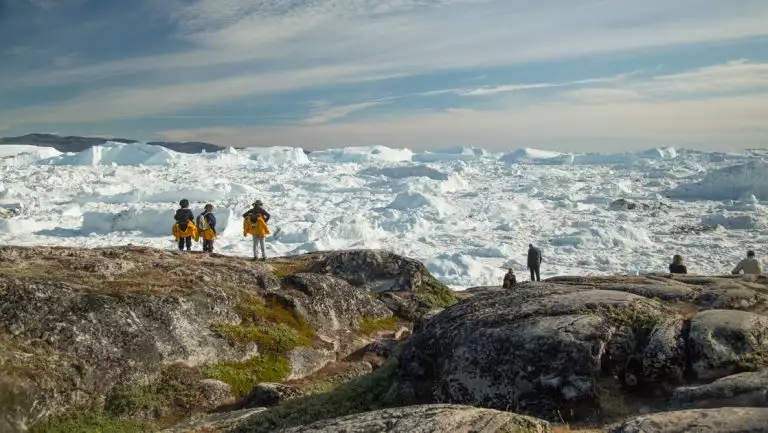 Hikers on a Northwest Passage small ship cruise stand at an overlook of tundra & rock looking onto an ice sheet in the sun.