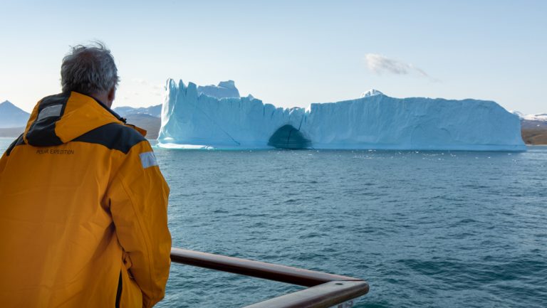 A Northwest Passage small ship cruise traveler in yellow jacket stands on deck of the boat, gazing at a large iceberg in the sun.