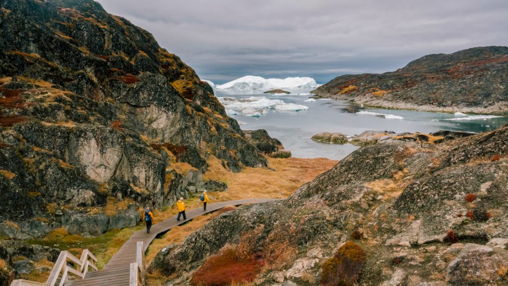 3 Northwest Passage small ship cruise travelers walk a wooden boardwalk towards the shore & icebergs, past rocks & tundra.