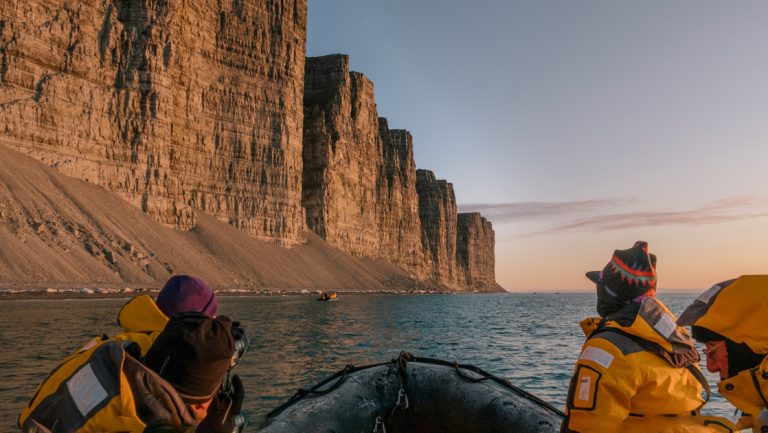 Northwest Passage small ship cruise travelers in yellow parkas ride in a Zodiac boat looking up at massive sandy cliffs at dusk.