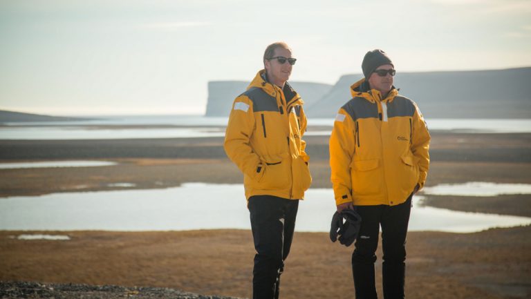 2 Northwest Passage small ship cruise passengers in yellow jackets, black pants & sunglasses stand among dusty rock & small ponds.