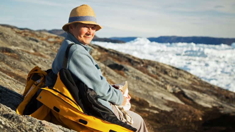 Woman in woven hat, blue sweater & yellow coat sits on rock face overlooking ice field in the distance on a sunny day.