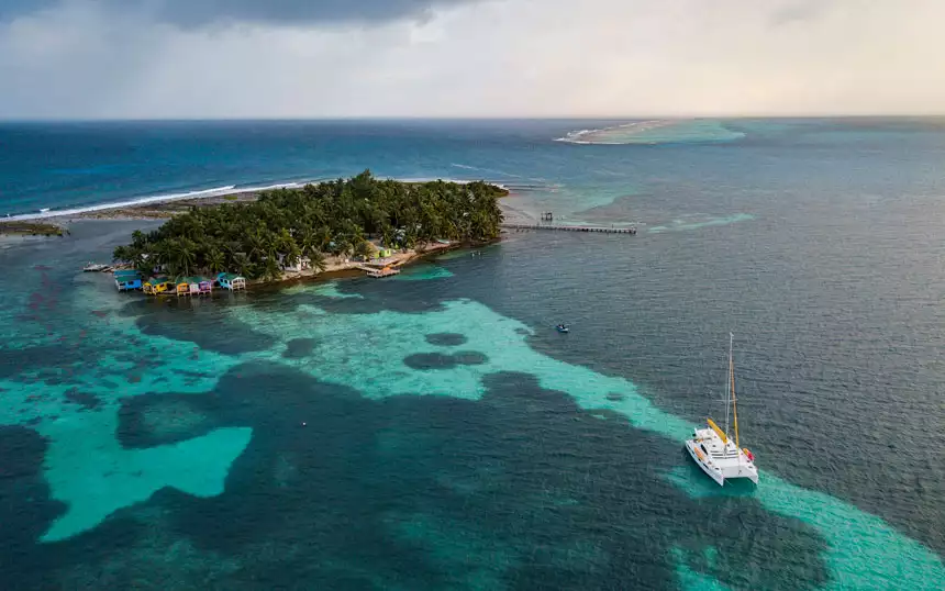 Aerial view of Tobacco Caye, surrounded by turquoise waters, reef & a private Belize catamaran charter yacht anchored near the island.
