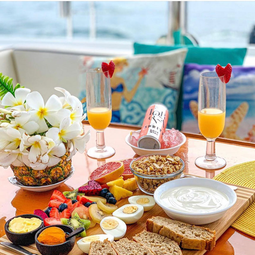 Tropical breakfast for 2 with mimosas, eggs, yogurt, fruits & bread set out on a sunny wood table aboard a charter boat in Belize.