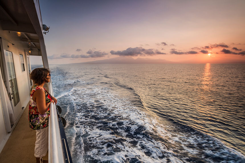 Woman stands on deck of small ship on romantic cruise in Hawaii, watching the sunset in calm seas.