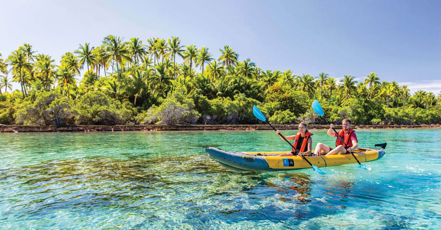 A girl and boy paddle a yellow kayak in crystal clear teal water front of a palm tree filled sandy island in Polynesia.