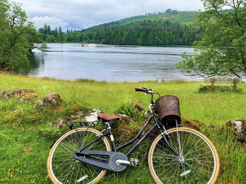 A bicycle with a wicker basket rests on the green grass alongside the Caledonian Canal, an activity option aboard Spirit of Scotland barge.