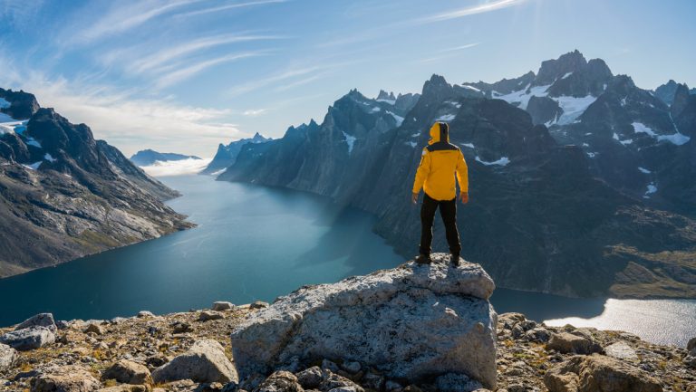 Greenland adventure cruise traveler stands atop mountain's edge looking down onto idyllic fjord among jagged mountains in sun.