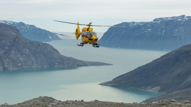 Yellow helicopter flies above large fjord with glacier & smooth mountains during a South Greenland adventure cruise.