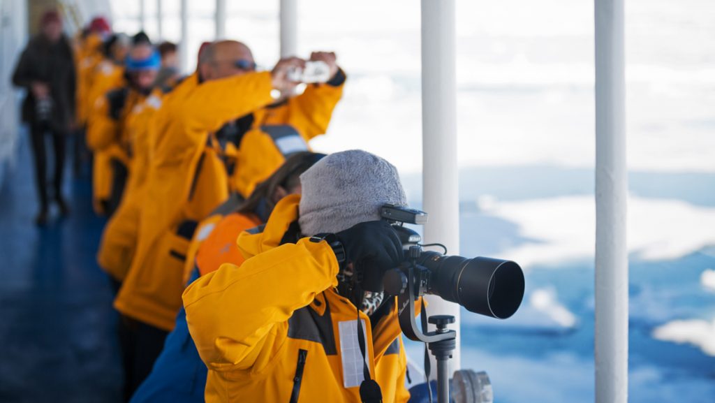 Arctic travelers in yellow coats stand on deck of ship photographing icebergs on the Spitsbergen Photography cruise.