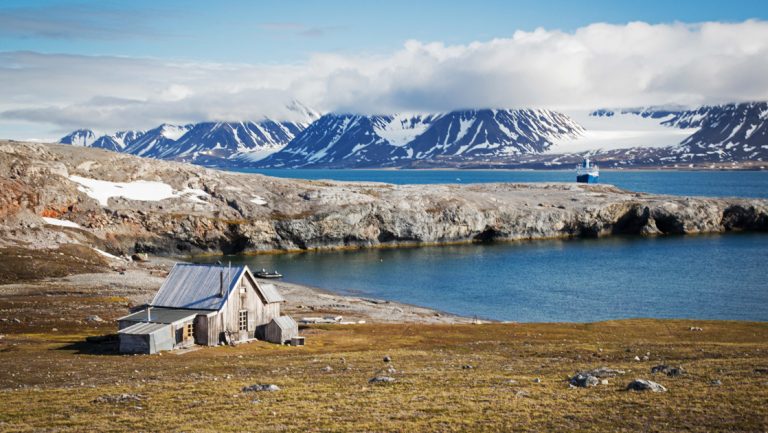 Old wooden abandoned trapper's hut sits on arctic tundra beside a fjord inlet with snowy mountains & small blue & white ship behind.