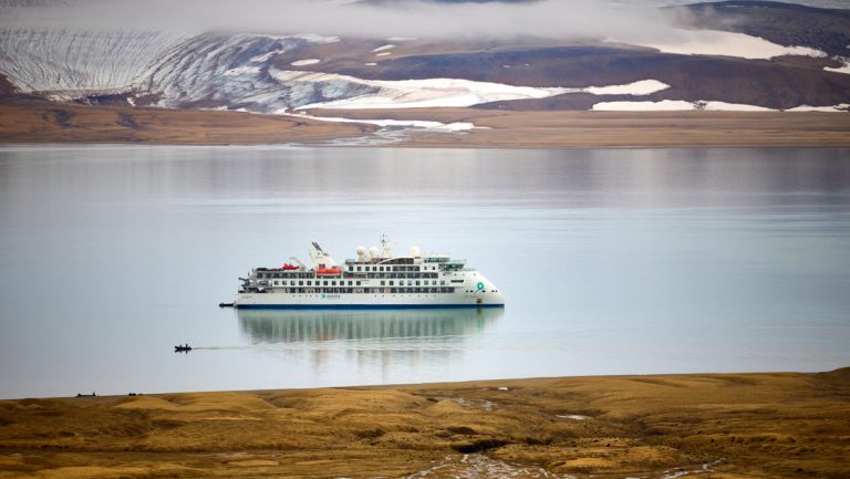 Small white expedition ship sits in glassy fjord while Zodiac cruises nearby with golden tundra on 1 shore & snow on the other.