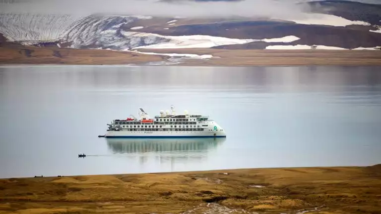 Small white expedition ship sits in glassy fjord while Zodiac cruises nearby with golden tundra on 1 shore & snow on the other.