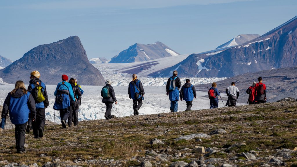 Line of Arctic Circle travelers walk over tundra looking down on large glacier with tall snow-capped mountains beyond.